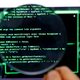Photo shows a magnifying glass over a screen with computer code.